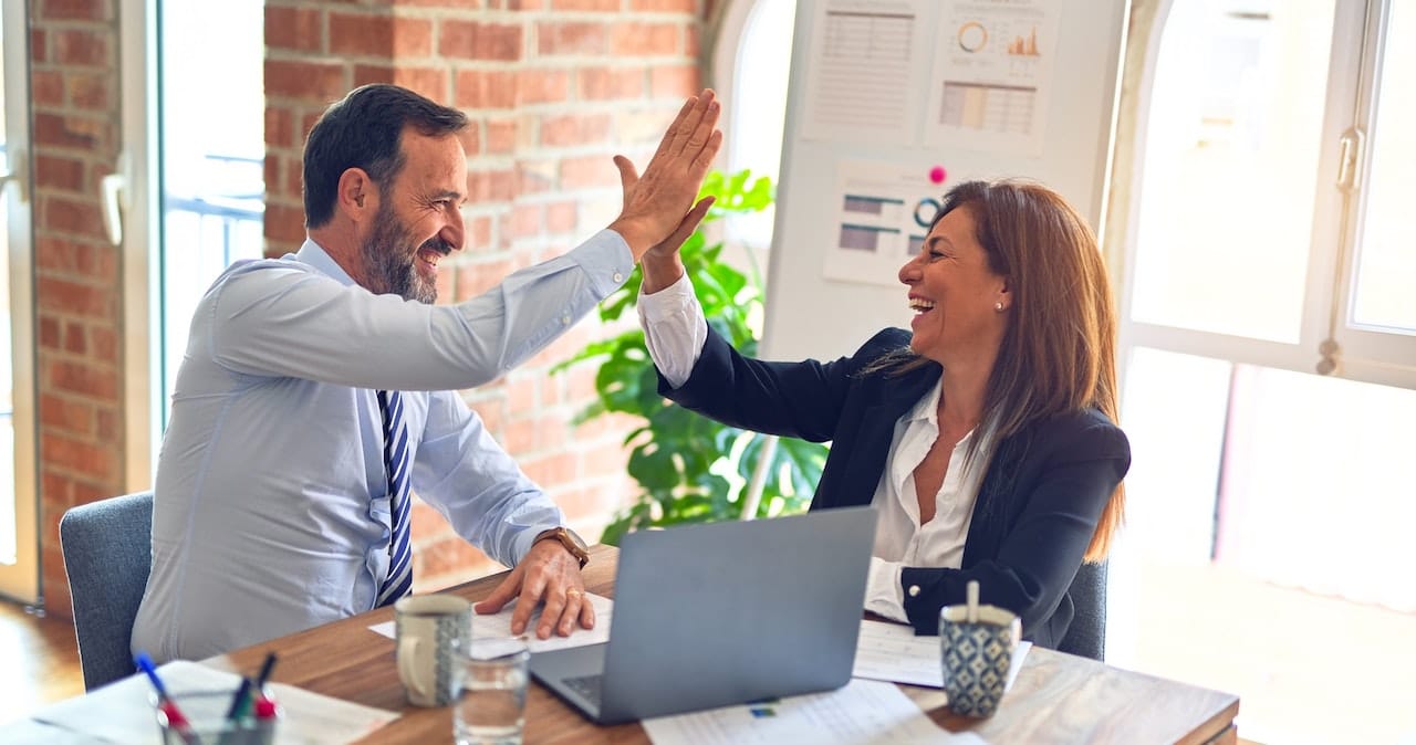 A man and woman sitting at a desk in an office giving each other a high five
