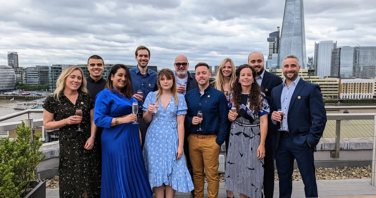The staff of Magenta Associates posing for a group photo on a rooftop bar with the London skyline in the background