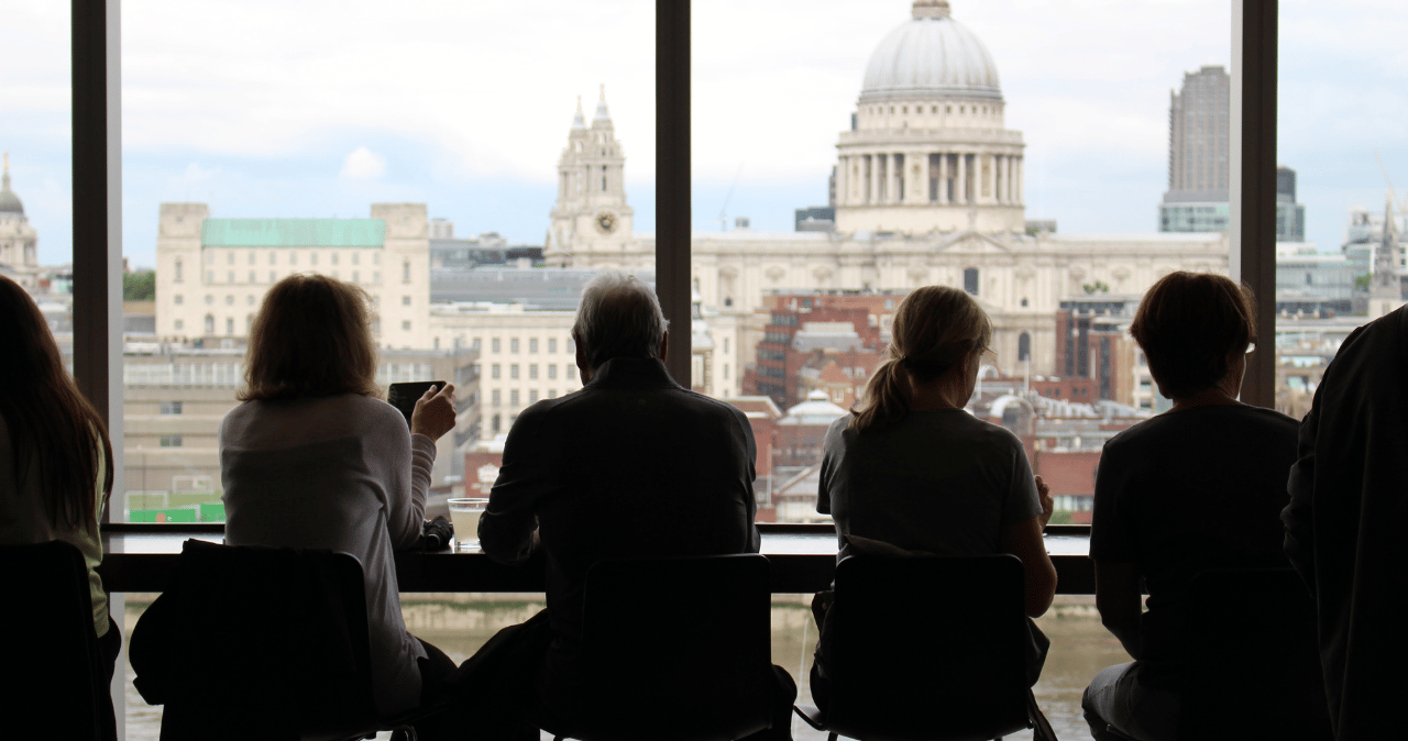 various people sit across from a window, outside the window is the landscape of London