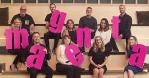 The employees of Magenta Associates sitting on three levels of steps, holding pink letters that spell out Magenta